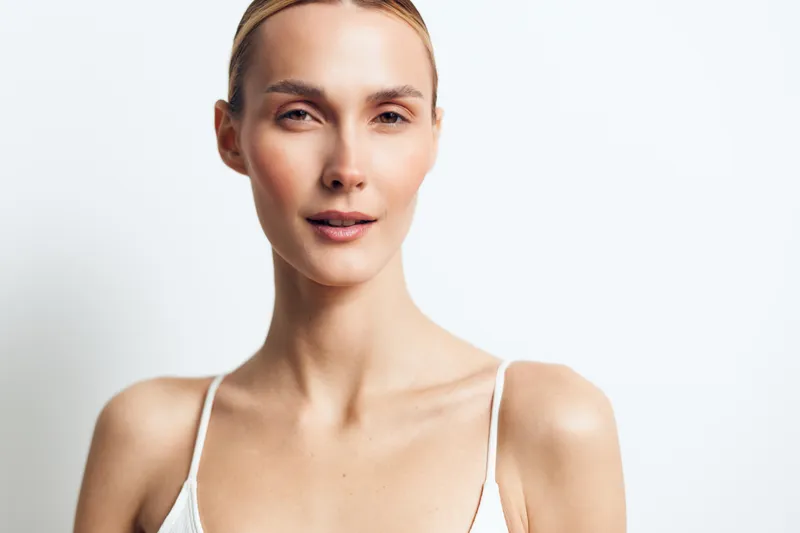 Young blonde woman with slicked back hair smiles softly wearing a white spaghetti strap top against a plain white background in a bright studio setting.