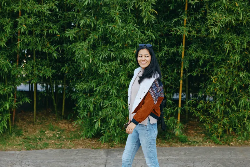 Woman, fashion, outdoor, casual, street, smile, bamboo, greenery, cardigan, denim, strolling beside lush plants in natural light, urban atmosphere, candid fashion moment