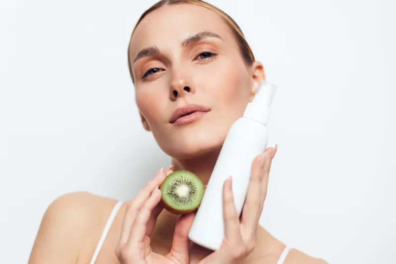 Close-up of woman holding a white spray bottle and a sliced kiwi fruit. Concept of natural skincare, beauty routine, and fresh ingredients. Soft lighting highlights clear skin and healthy glow.