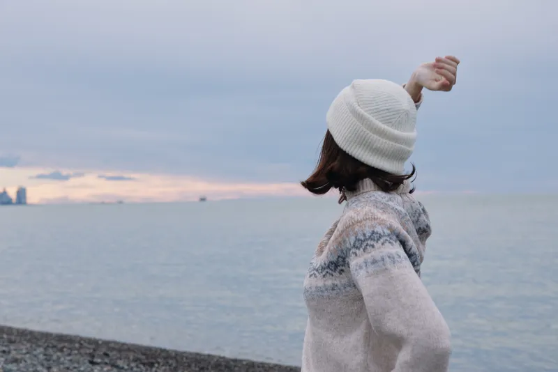 Person on a quiet beach facing the sea in a cozy sweater and beanie, a moment of relaxation with calm horizon and winter mood by the water.