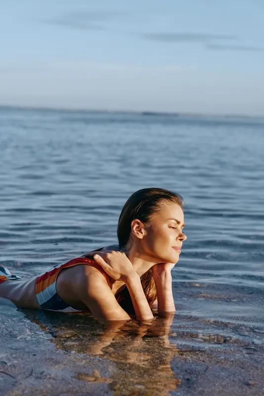 Woman beach ocean waves swimming relaxation nature health fitness summer, young athlete in shallow seawater at shoreline, calm morning sunlight, lying on elbows, profile view, peaceful mood