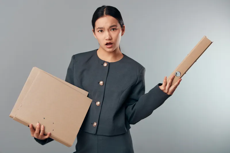 Woman holding cardboard boxes with confused expression wearing formal jacket. Delivery, packaging, logistics and shipping concept with uncertain female in gray outfit on plain background.