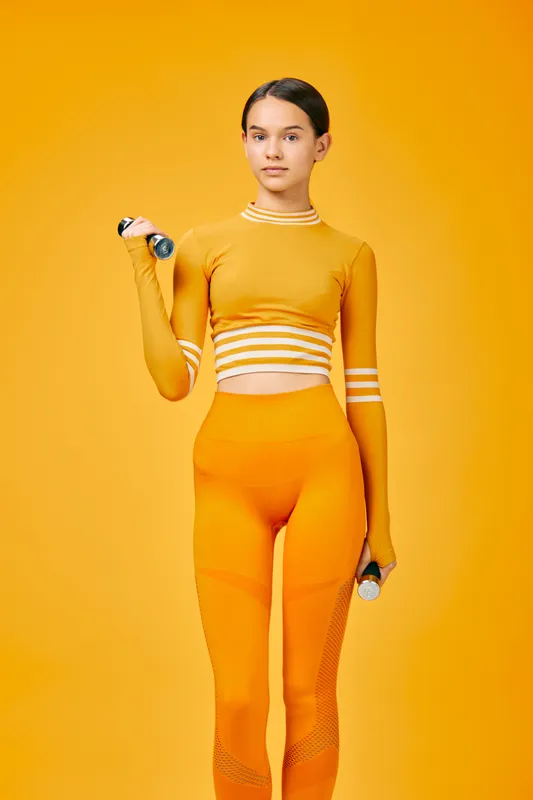 Fitness woman in bright orange athletic wear stands against a vivid orange backdrop, holding dumbbells ready for a training session, showcasing strength, balance and focused determination in a