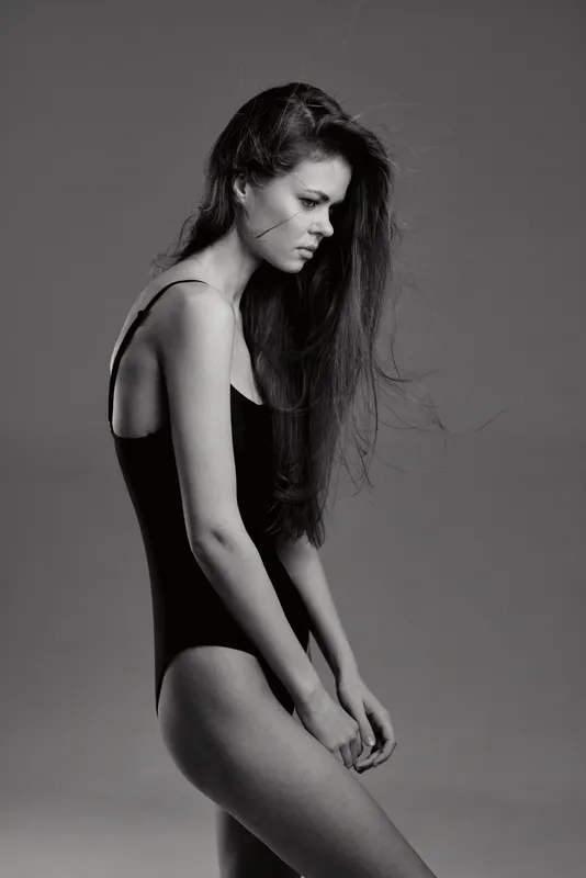 Black and white portrait of a thoughtful woman in a black bodysuit with long hair against a plain background.