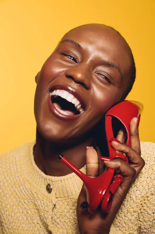 Happy African woman with dark skin laughs joyfully holding a red high heel shoe near her face on yellow background. Concept of fashion, beauty, and positive emotions in studio.