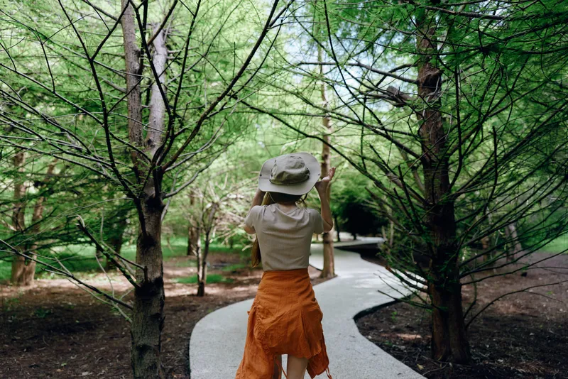 Woman walking on winding path through forest wearing hat and orange dress, nature trail with trees and dappled sunlight, casual summer stroll and peaceful outdoor exploration.