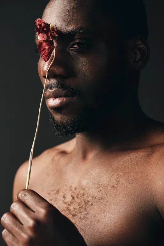Creative conceptual portrait of African man with dark skin holding dried flower near face in visual artistic style. Closeup, bare chest, moody lighting and emotional expression for beauty and