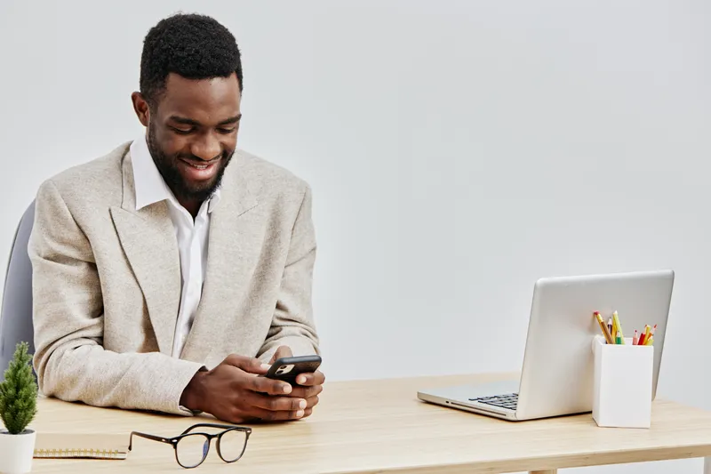 Smiling man using smartphone at desk with laptop and stationery, casual office environment, light background, relaxed mood, modern workspace, young adult male, technology, communication, freelance wor