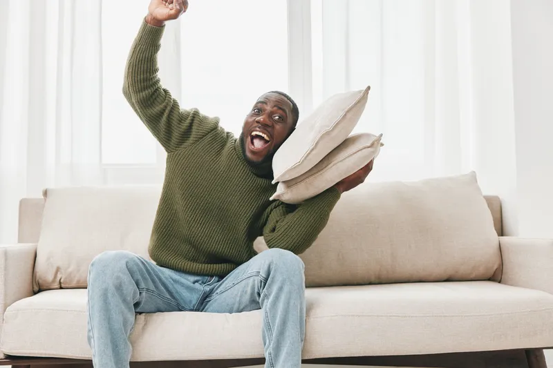Joyful African man celebrating at home, holding pillows in a cozy living room, wearing a green sweater and denim jeans, conveying happiness and excitement