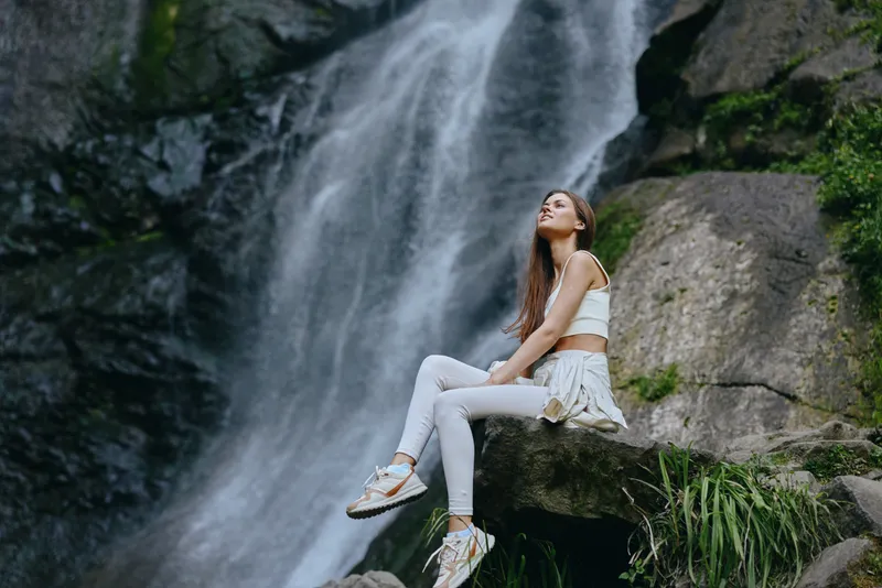 Young woman sitting on rocks near a waterfall, wearing white activewear, enjoying nature and calm surroundings in lush green forest outdoors.