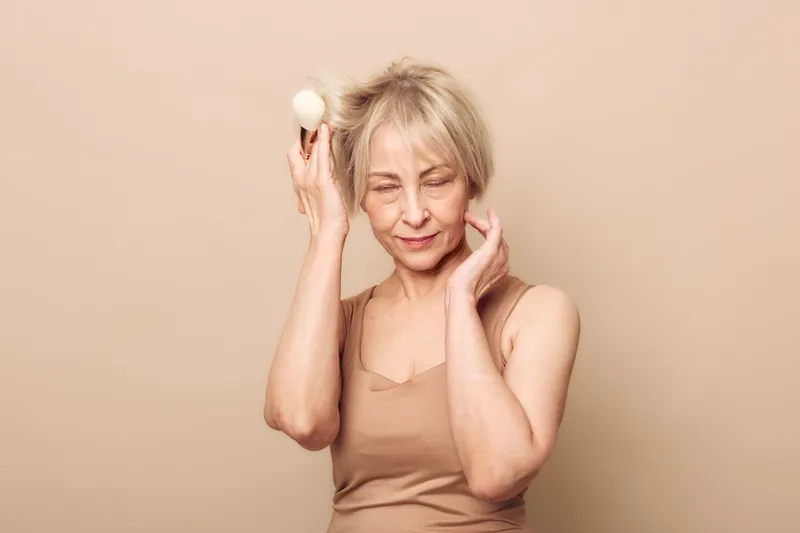 Blonde middle aged woman with short hair holds white facial cleansing brush near face, eyes closed, wearing beige tank top. Concept of skincare, self care, beauty, and natural routine.