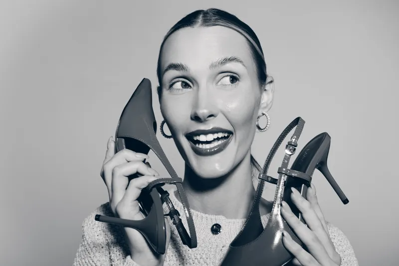 Young white woman with light skin smiling and holding red high heel shoes near face, showing happiness and fashion concept on gray background.
