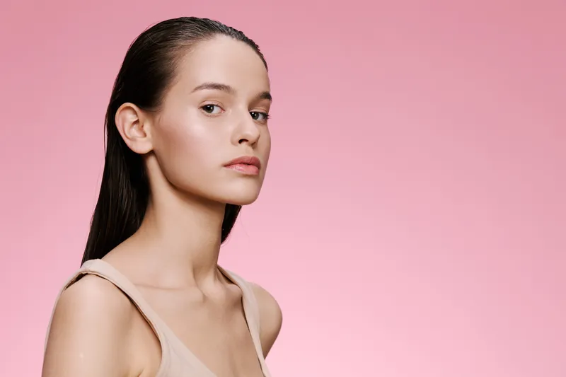 Portrait of a young model with natural beauty and minimal makeup against a soft pink background, symbolizing freshness and simplicity