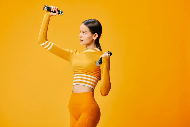 A woman fitness enthusiast lifts dumbbells in a bold orange studio, showcasing a dynamic workout pose, strength and vibrant athletic style for modern sport imagery and energy.