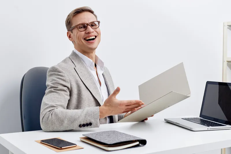 Smiling middle-aged man in business suit sitting at office desk holding a document, casual working posture, cheerful expression, modern workspace with laptop, notepad and smartphone, professional envi