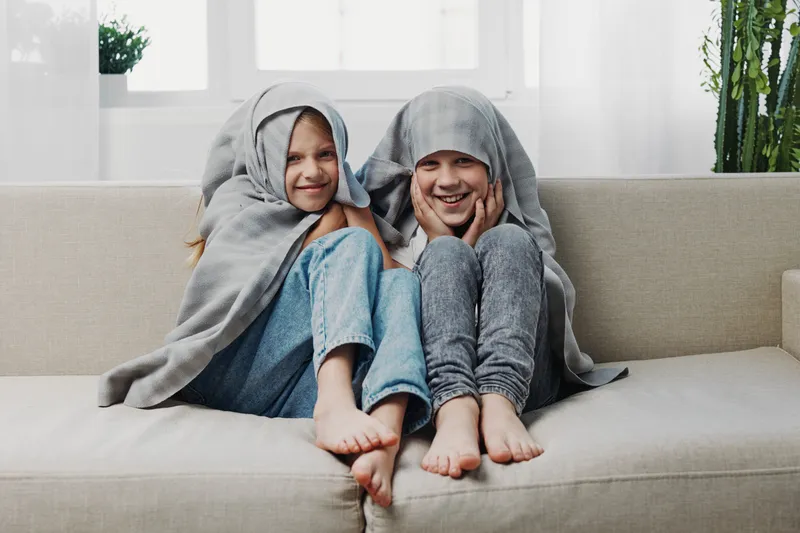 Two happy children wrapped in gray blankets sit barefoot on a beige sofa, smiling and enjoying a cozy moment in a bright living room with plants in the background.