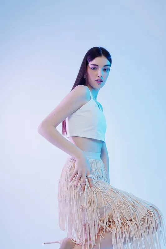 Young woman in a white crop top and fringed skirt poses elegantly against a light background. Concept of beauty, fashion, and modern style in studio lighting.