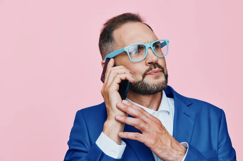 A business professional man on a phone, expressing thoughtful emotion against an isolated pink background, captured in a studio shot that emphasizes modern work life and focus.