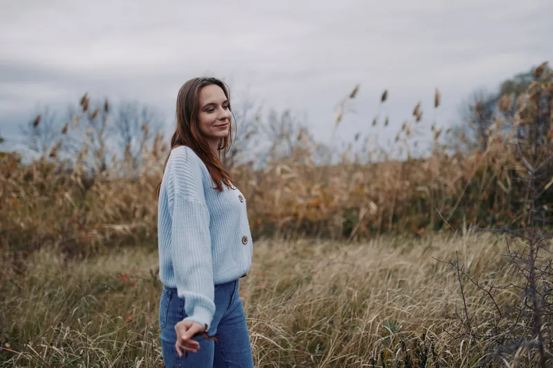 A young woman stands in a windswept field wearing a cozy sweater, natural pose and soft smile conveying authenticity and credibility in a candid outdoor portrait with muted tones.