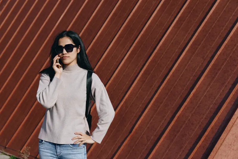 Casual street style with a sunglass clad woman on her phone, wearing a knit sweater and backpack against a striped wooden wall, urban fashion moment