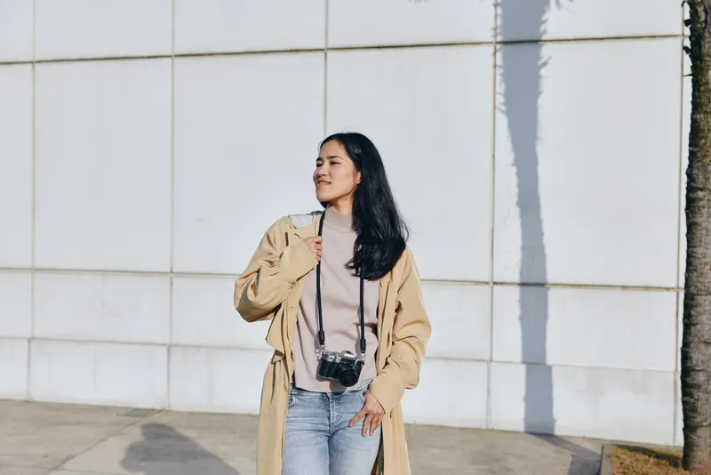 Woman with camera strapped at her neck, wearing a beige long coat and jeans, stands against a white tiled wall under sunlight, capturing a calm street moment with confident style.