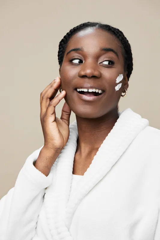 Cheerful young Black woman applying skincare cream on her face, wearing a white robe, against a soft beige background, representing self care and beauty