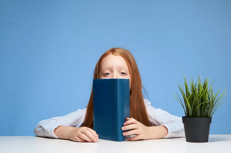 Рыжеволосая девушка сидит за школьной партой с книгой в руках