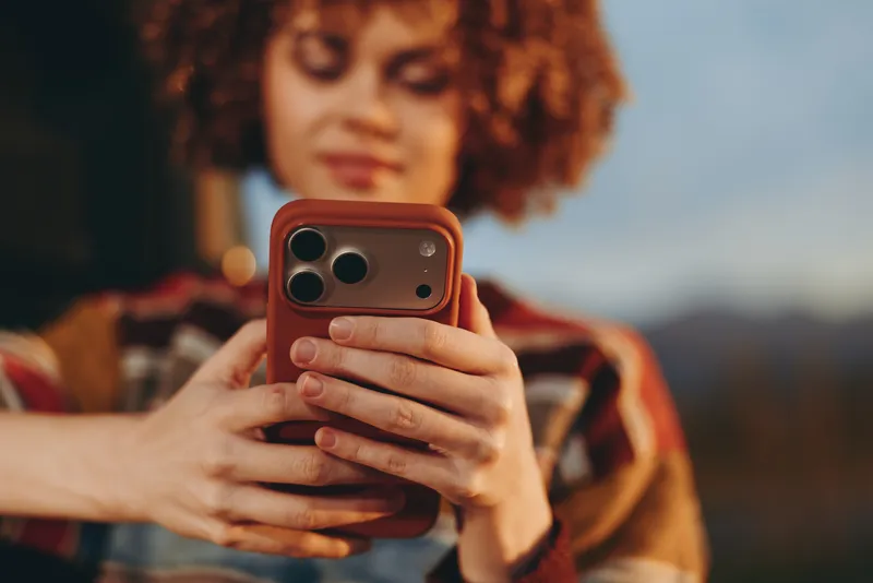 Woman lifestyle, holding smartphone for selfie in mirror view outdoors at sunset. Curly haired woman with warm evening light and blurred landscape background, casual portrait, soft focus.