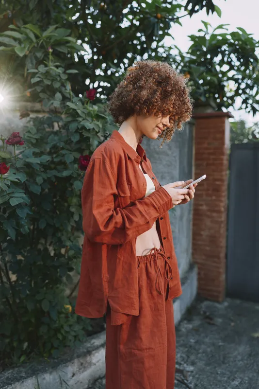 Young woman with curly hair wears a rust colored outfit and glances at her phone in a sunlit outdoor garden scene