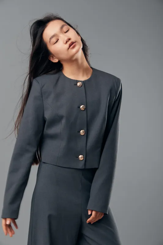 Woman in gray suit with buttons posing with closed eyes and relaxed expression against gray background. Female model with long dark hair wearing elegant stylish outfit in studio environment.