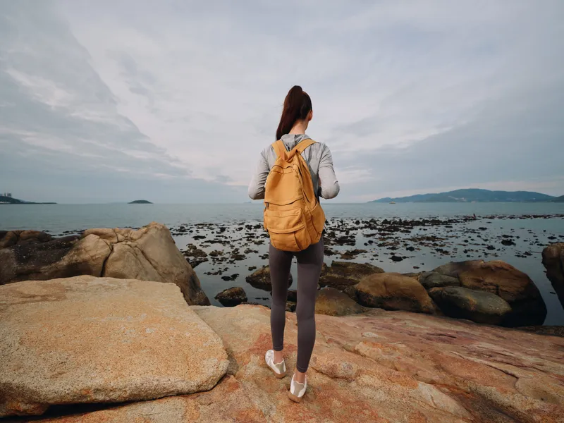 Young woman enjoying nature by the sea, wearing a backpack and activewear, exploring the rocky shore at dawn with a serene expression and a soft color palette