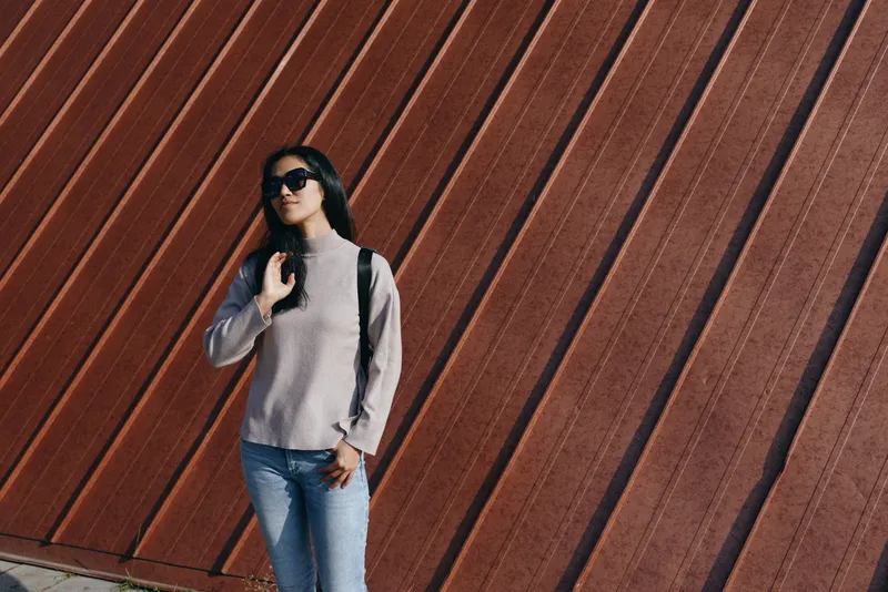 Model in casual street fashion posing against an angled wall, wearing sunglasses, jeans and backpack, outdoors in urban light and texture, capturing contemporary style and confident stance