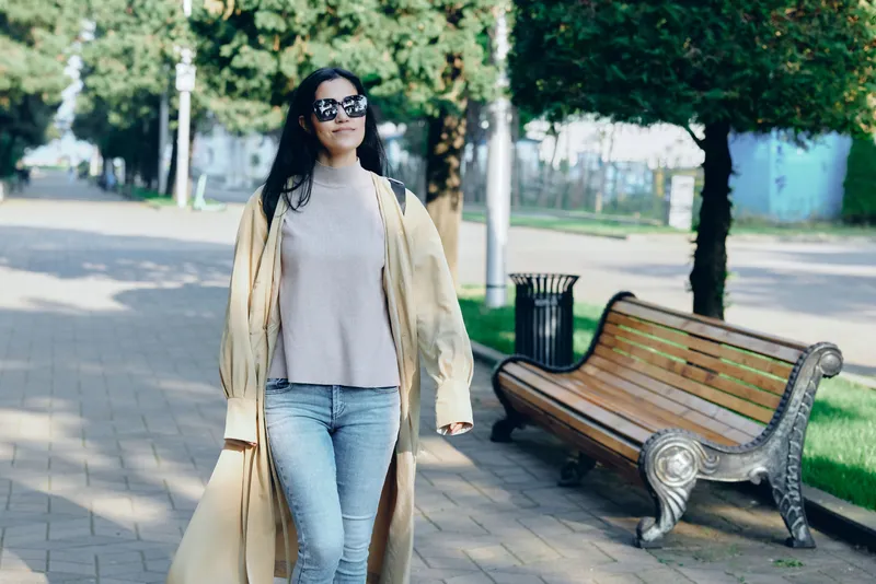 Woman wearing a beige coat and sunglasses walks along a sunny park path, showcasing casual chic fashion in an urban setting in daylight with relaxed vibe