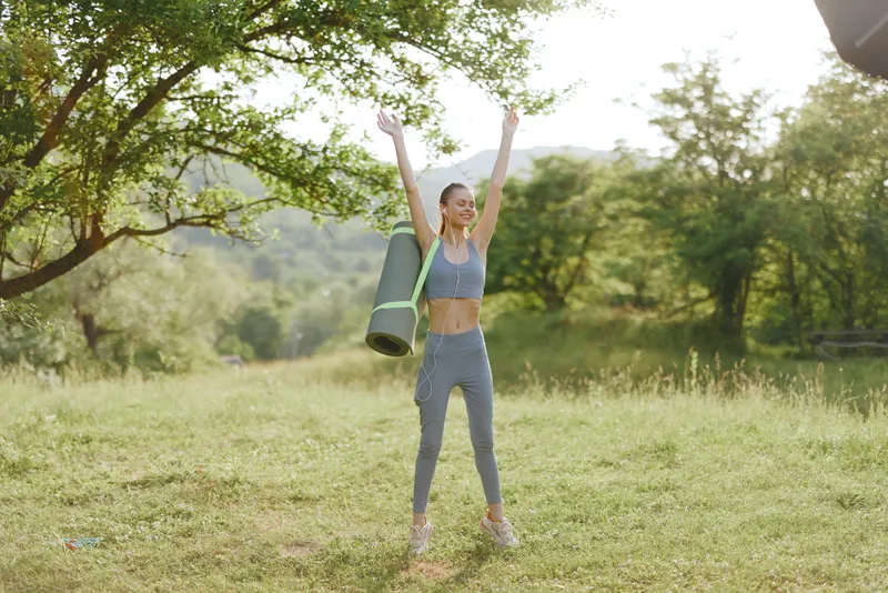 Energetic young woman celebrating outdoor fitness, wearing workout attire, enjoying nature, with a yoga mat, conveying health and vitality in a serene environment