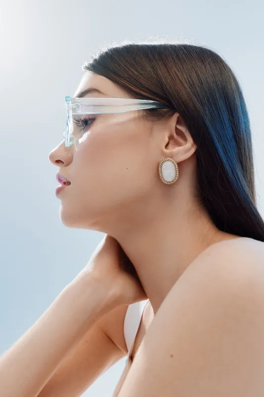 Young woman with long dark hair wearing transparent glasses and elegant earrings poses in profile on a light background. Concept of beauty fashion and style.