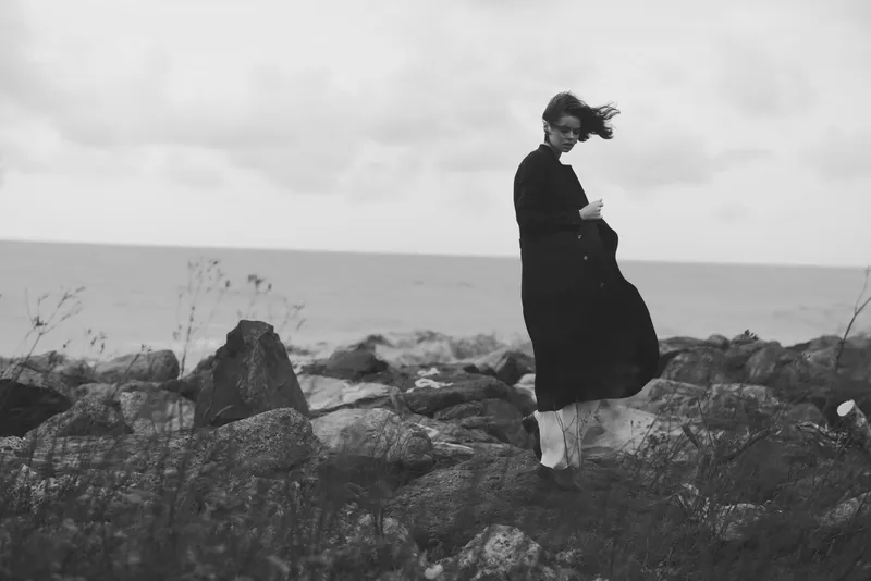 Monochrome scene of a windswept woman standing on rugged rocks beside the sea, cloak fluttering, contemplative mood, distant horizon, solitary figure against expansive coastline, minimalist