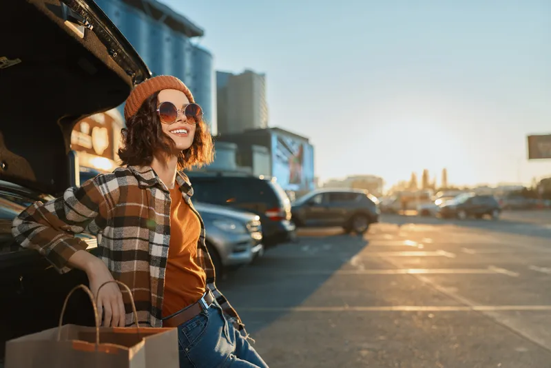 Woman by car trunk in urban parking at sunset, smiling with shopping bags; candid lifestyle portrait emphasizing authenticity, golden hour glow, mindful living and emotional storytelling.