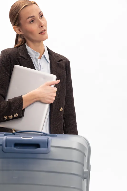 Young businesswoman in formal wear holding laptop and standing next to suitcase. Concept of travel, business trip, preparation, and modern professional lifestyle.