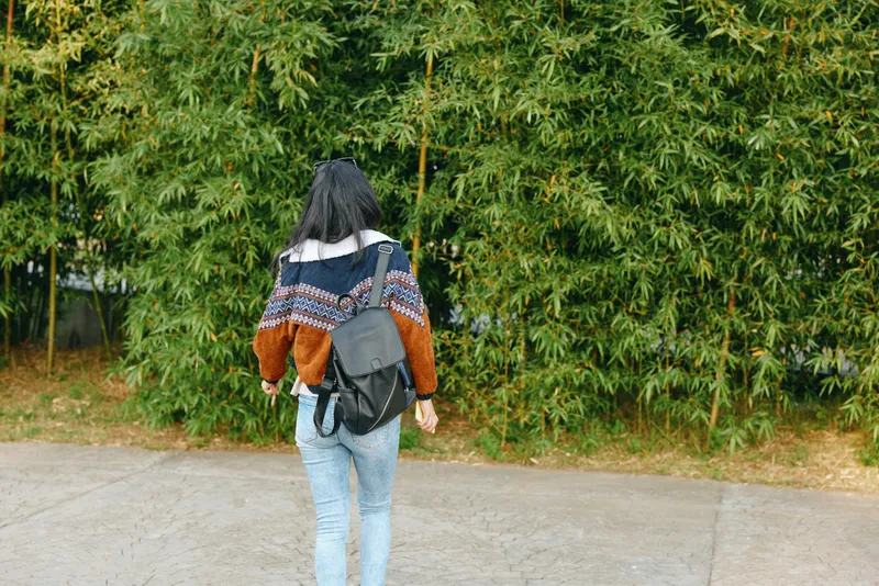Back view of a woman walking along a sidewalk beside dense greenery, wearing a patterned sweater, light jeans and a backpack, capturing casual street style and outdoor exploration.