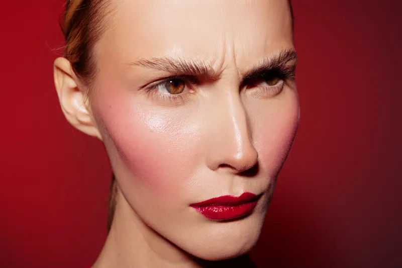 Close-up of a woman with red lipstick and flushed cheeks frowning against a red background. Concept of emotion, beauty, makeup, and facial expression in studio lighting.