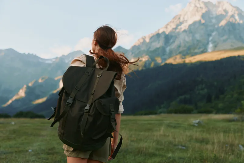 Young woman with a backpack hikes toward distant mountains, feeling adventurous and free on a sunny outdoor day.