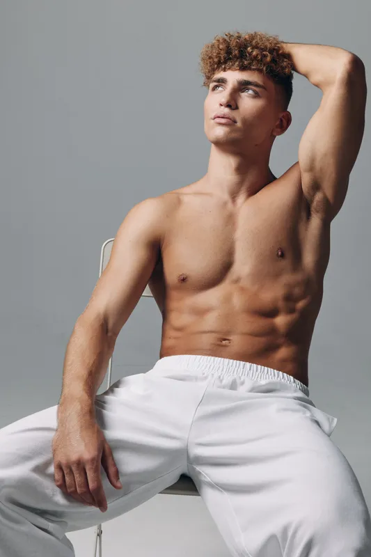 Young man with muscular body and curly hair posing shirtless in white pants against gray background. Fitness, confidence and casual style with relaxed posture and thoughtful expression.