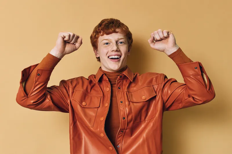 Redhead young man with freckles wearing brown leather jacket and dark shirt smiling and raising arms in excitement against beige background in studio portrait.