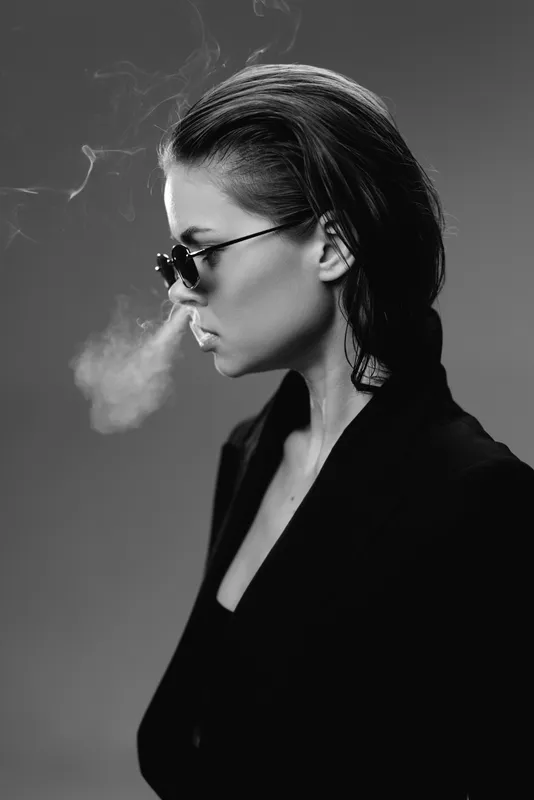 Black and white portrait of a smoking woman in sunglasses with slicked-back hair and dark clothing, exuding a moody, stylish vibe.