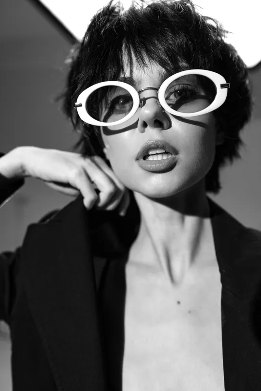 Black and white portrait of a fashionable woman wearing oversized white sunglasses and dark blazer with confident expression.