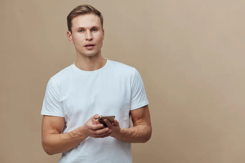 young caucasian man holding smartphone wearing white t-shirt standing beige background digital nomad technology communication concept