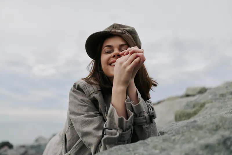 Young woman wearing a checkered coat and matching cap smiles with hands clasped near her face outdoors by rocky shore. Overcast sky creates soft natural lighting and relaxed mood.