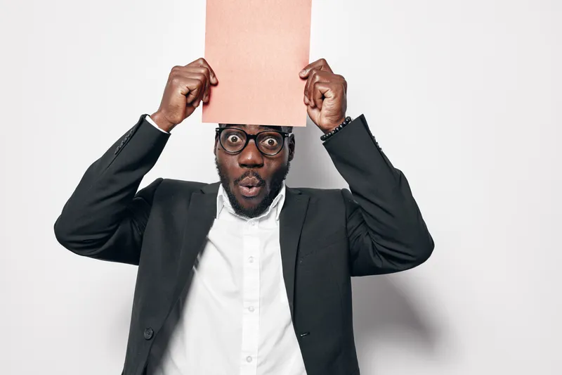 African man with dark skin and glasses holding blank paper above head with surprised expression in studio. Black male in suit and white shirt on white background with copy space.