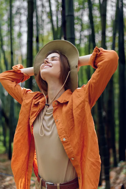 Woman in hat in forest with smile outdoors among bamboo, relaxed portrait showing nature and orange jacket, casual fashion style enjoying peaceful relaxation, happy serene mood and fresh air.