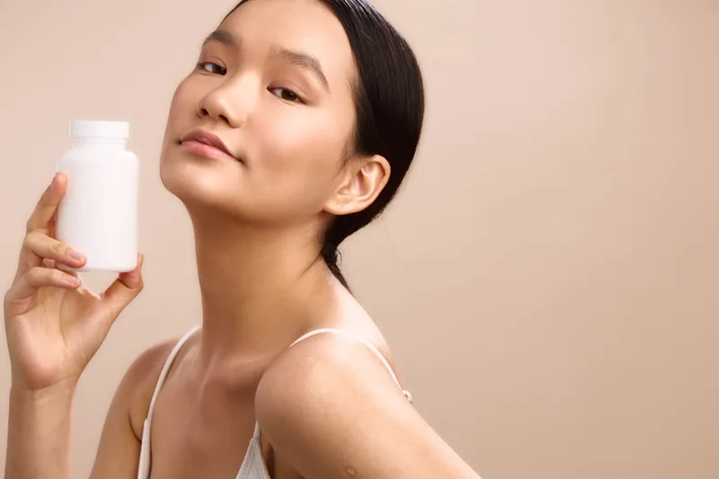 asian woman holds a white bottle near her shoulder, smiling softly in a minimalist studio, showcasing skincare and a fresh, confident beauty moment in a clean, bright setting.